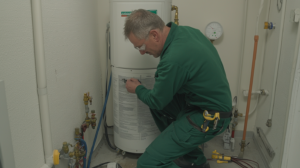 Plumber in a green uniform repairing a cylindrical white water heater in a utility room with visible pipes and a pressure gauge.